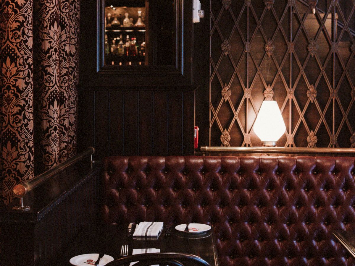 A cozy dimly lit restaurant corner with a burgundy tufted leather bench, a small table set for two, and an elegant lamp providing warm light.